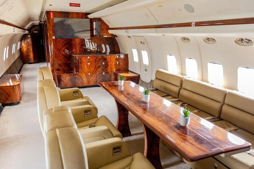 A long polished wood dining table and cream seating in a luxurious aircraft cabin.