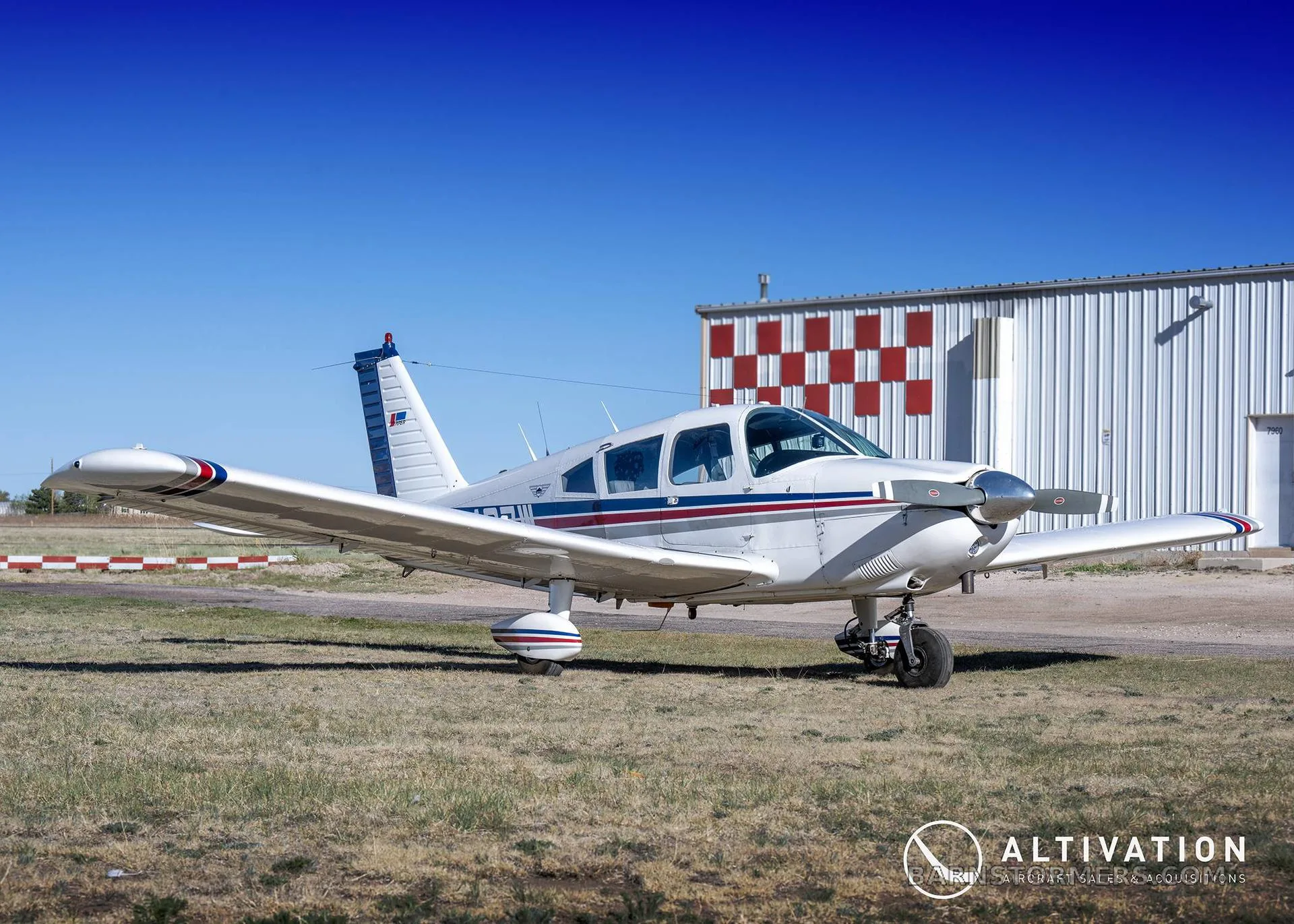 A white single-engine propeller aircraft with blue and red stripes is parked on grass next to a hangar under a clear blue sky.