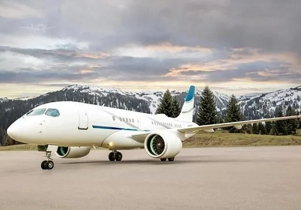A white Embraer business jet with blue accents on the tail and engines, parked on an airfield with snow-capped mountains in the background under a cloudy sky.