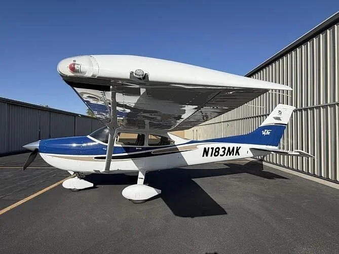 A blue and white high-wing single-engine aircraft with registration N183MK is parked on an asphalt ramp next to a hangar.