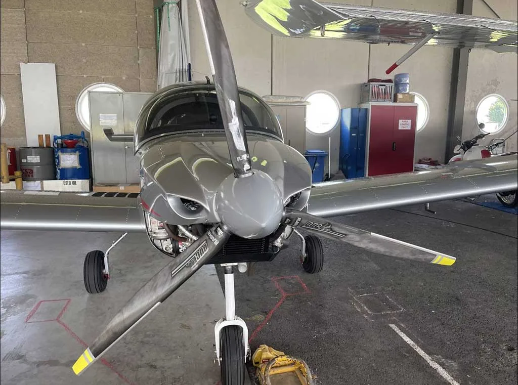 Head-on view of a silver Sonaca 200 aircraft with a three-bladed propeller, parked in a hangar.