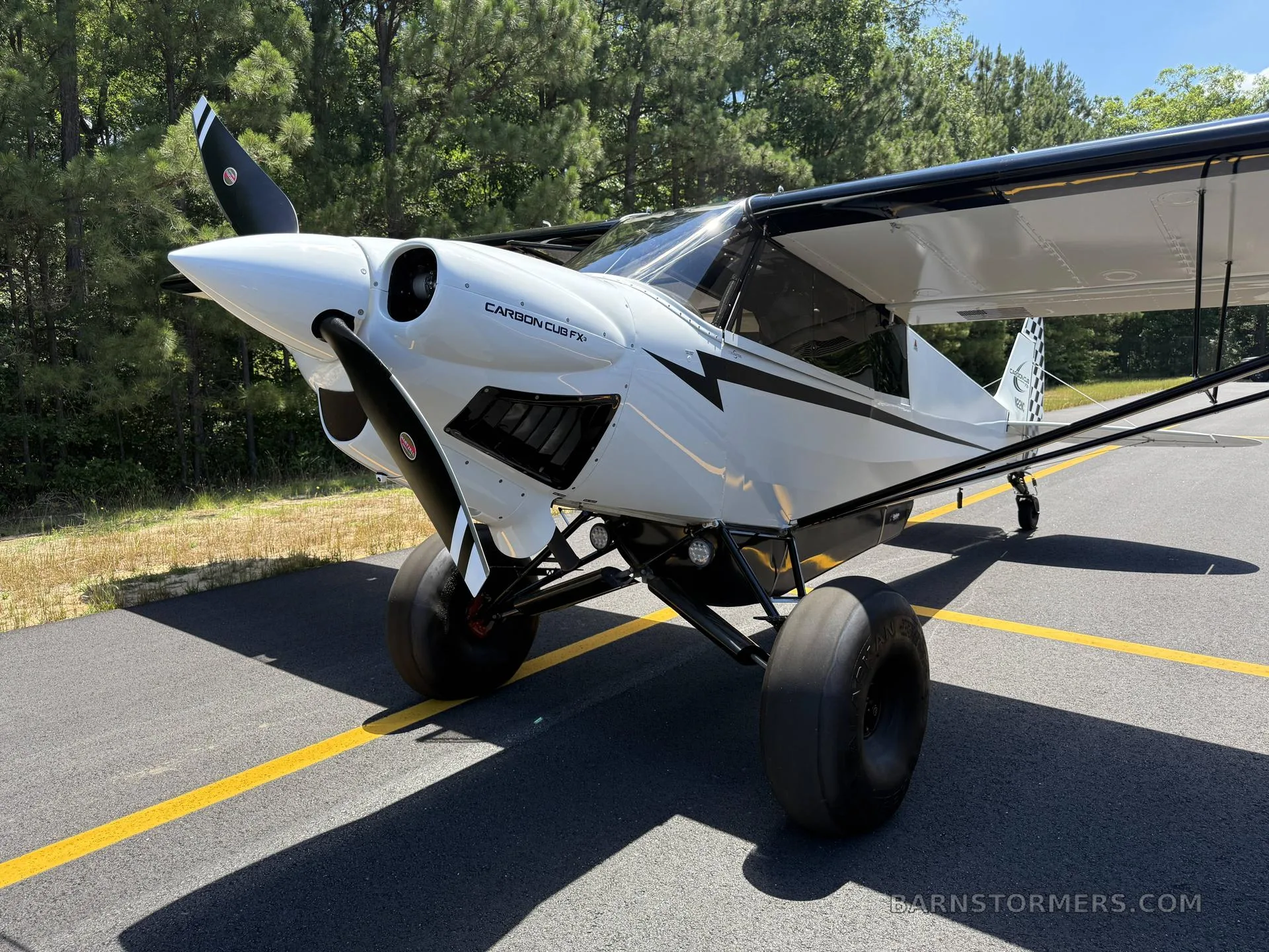 A close-up front-quarter view of a white Carbon Cub FX-3 aircraft with black stripes and large tundra tires is parked on an asphalt runway.