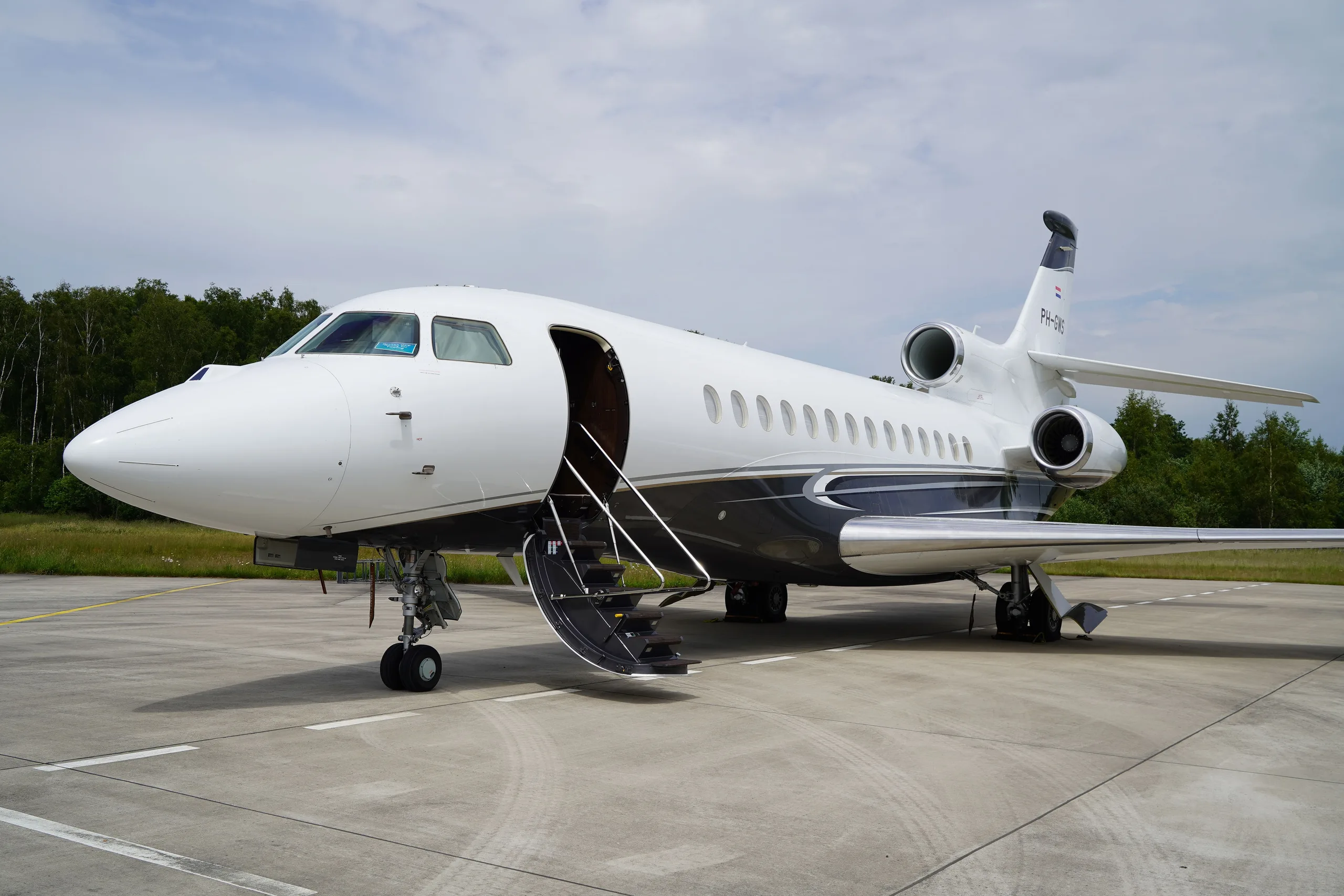 A white Dassault Falcon business jet with dark grey and silver accents is parked on an airfield with its main door open.