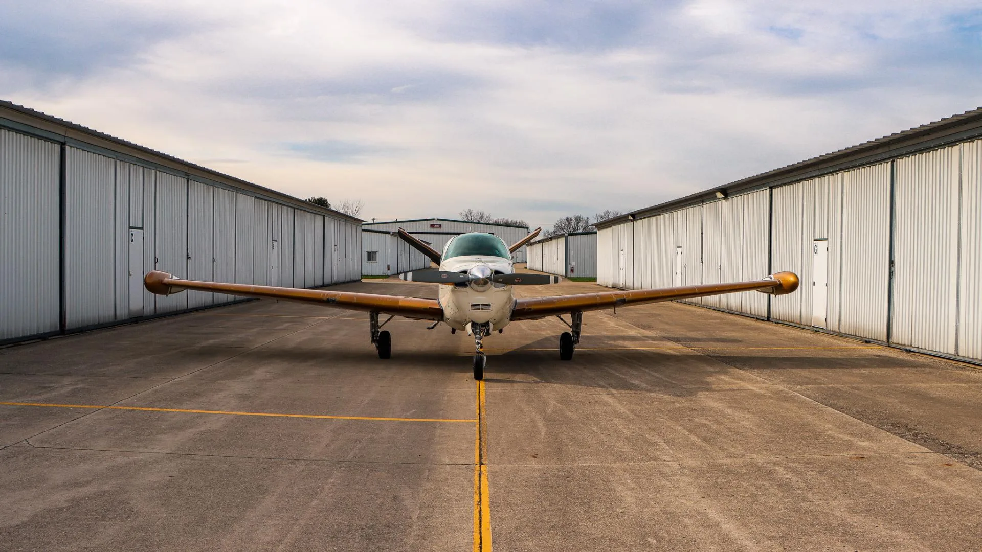 Front view of a white and gold Beechcraft Bonanza parked on an airfield between hangars.