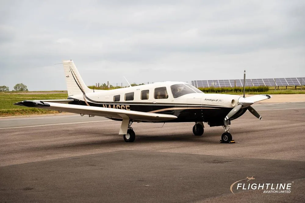 A white and black Piper Saratoga II TC, registration N446SE, parked on an asphalt runway with solar panels in the background.