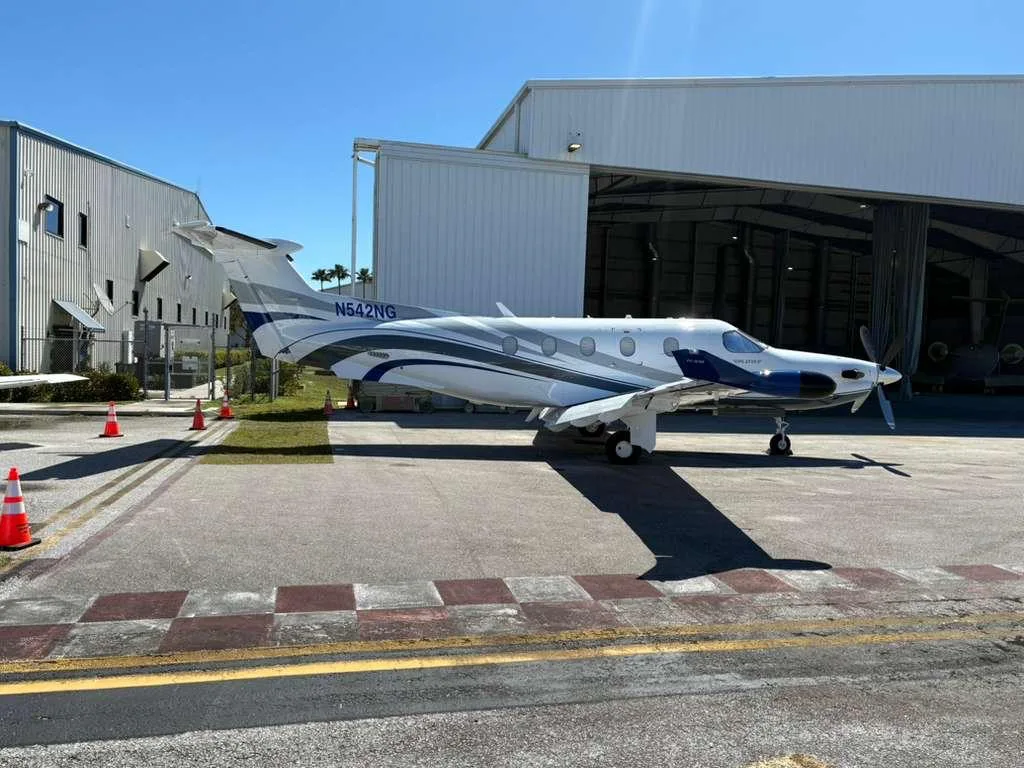 A white and blue Pilatus PC-12 NG aircraft with registration N642NG is parked on a tarmac next to hangars under a clear sky.