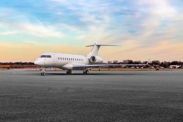 A white Bombardier Global business jet is parked on an airport tarmac under a colorful sky at dusk.