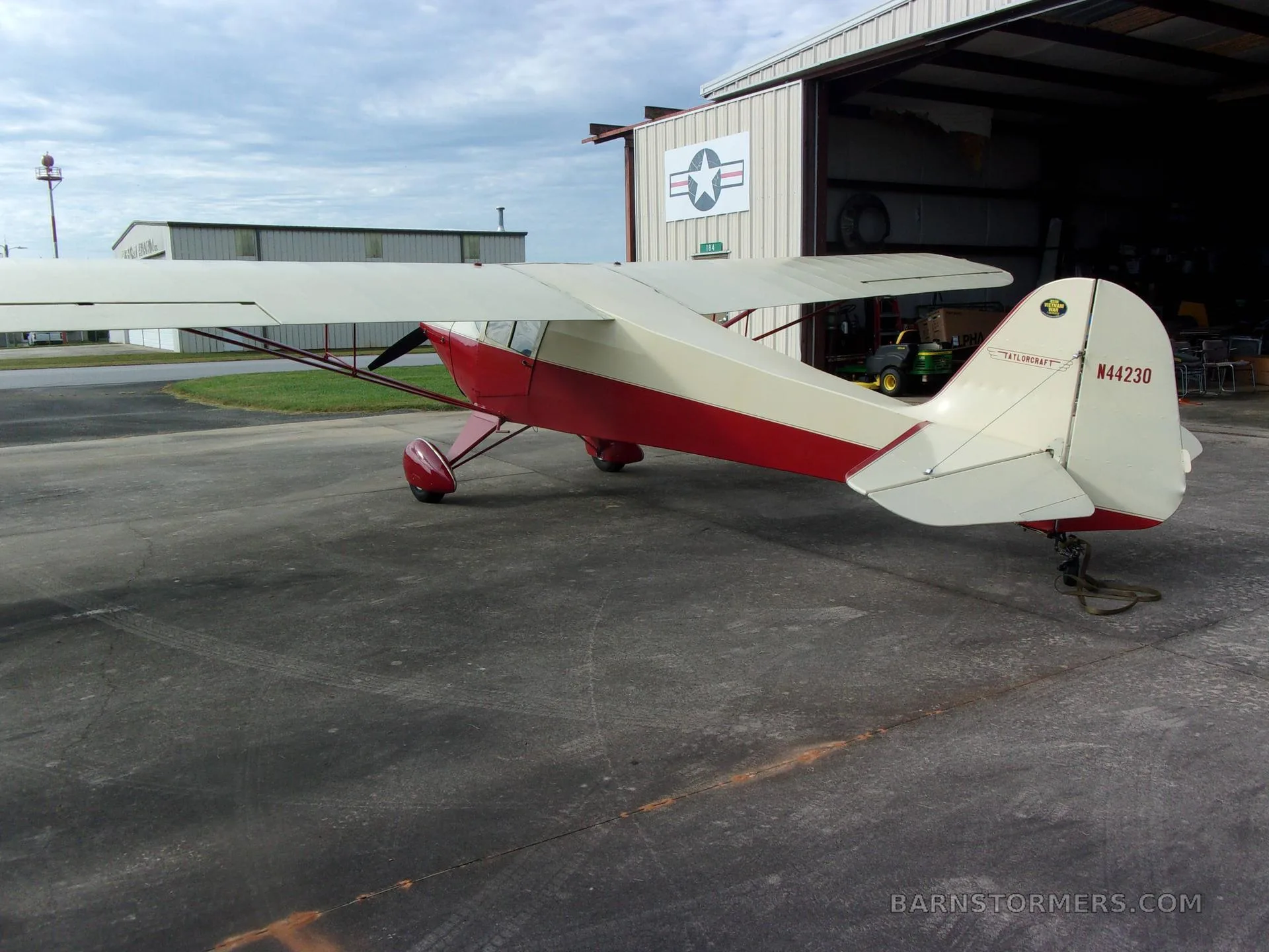 A red and cream Taylorcraft BC-12D aircraft with tail number N44230 parked on a concrete apron outside a hangar, viewed from the rear-right.