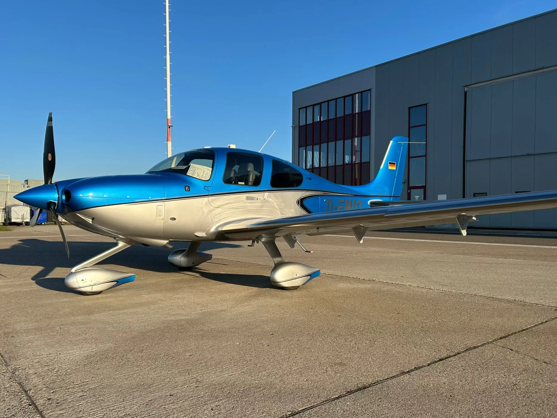 A blue and silver Cirrus SR22 aircraft parked on an asphalt apron in front of a hangar.
