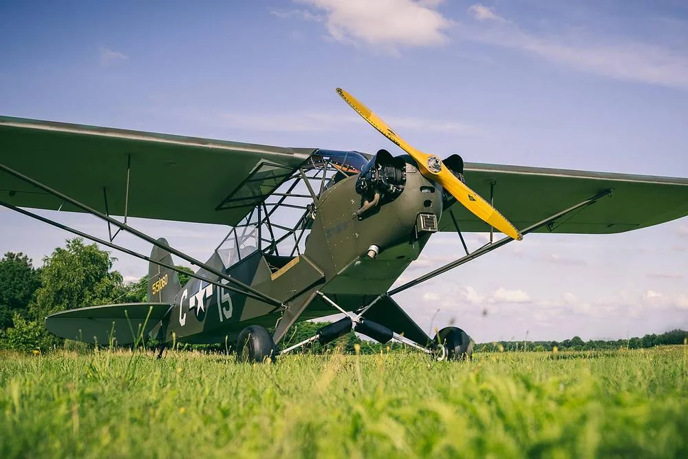 A low-angle front view of a Piper L-4 Grasshopper in military olive drab paint, with a yellow propeller, parked on a grassy field.
