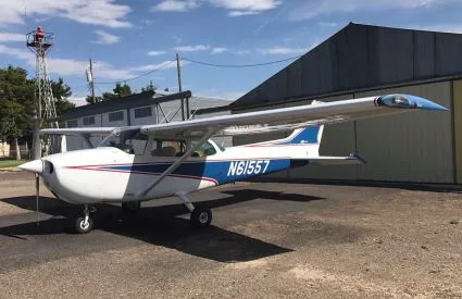 A white single-engine propeller aircraft with blue and red stripes, registration N61557, parked on an airfield in front of a hangar.