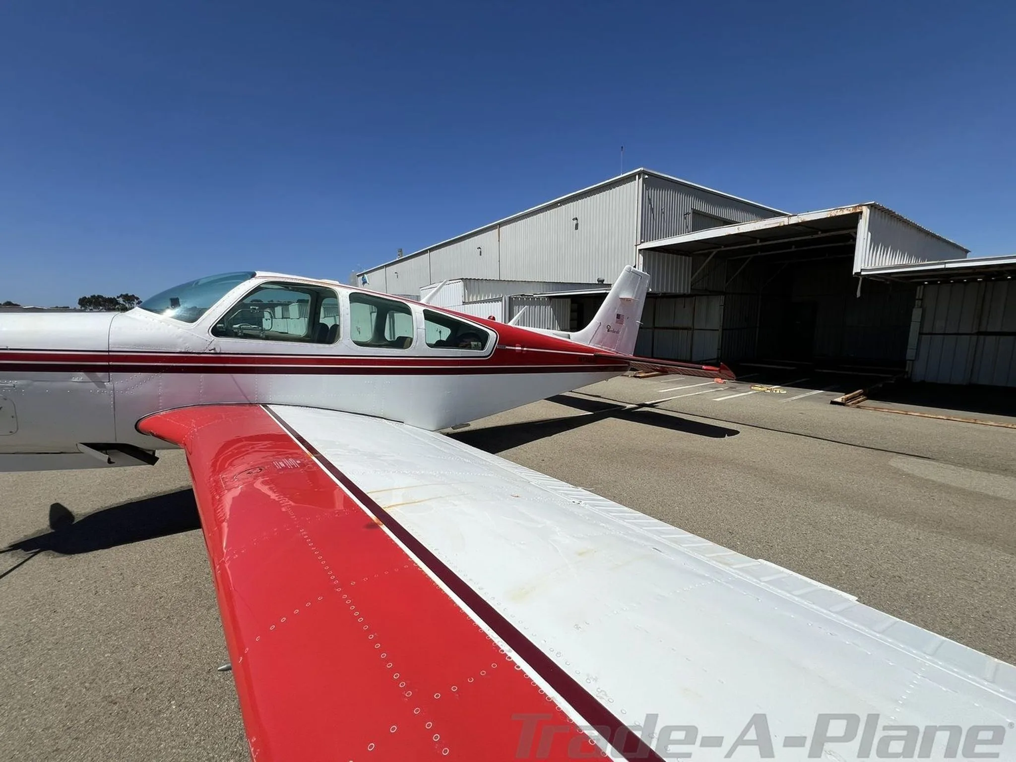 Rear-side view of a white and red Beechcraft Bonanza showing the V-tail and wing, parked near a hangar.