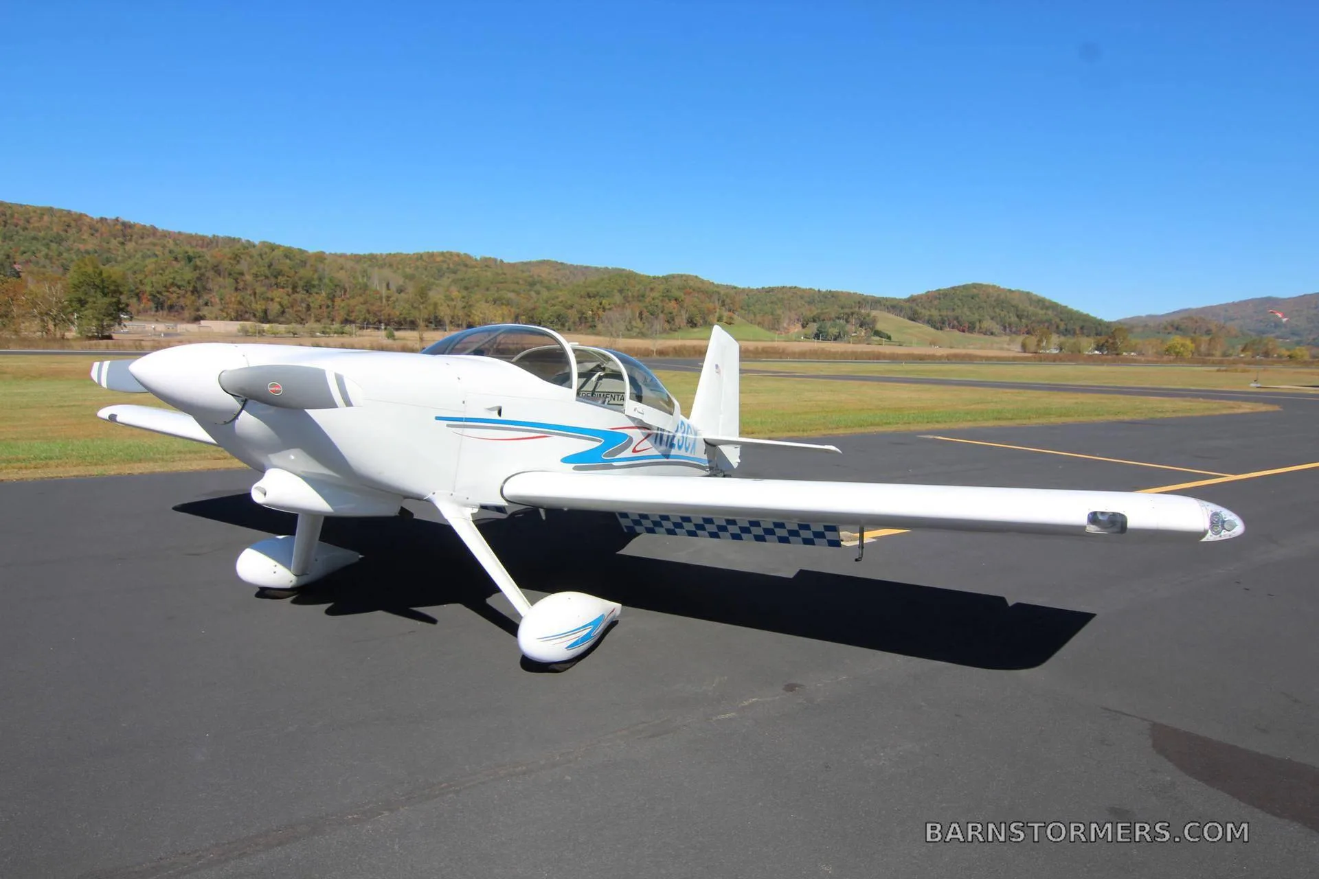 A white single-engine aircraft with blue and red stripes (N123CX) parked on an airfield under a clear blue sky with autumn hills in the background.