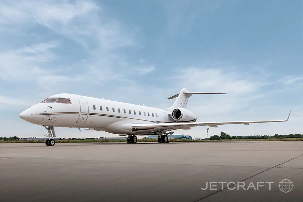 A white Bombardier Global business jet with gold and black stripes parked on an airfield under a partly cloudy sky.