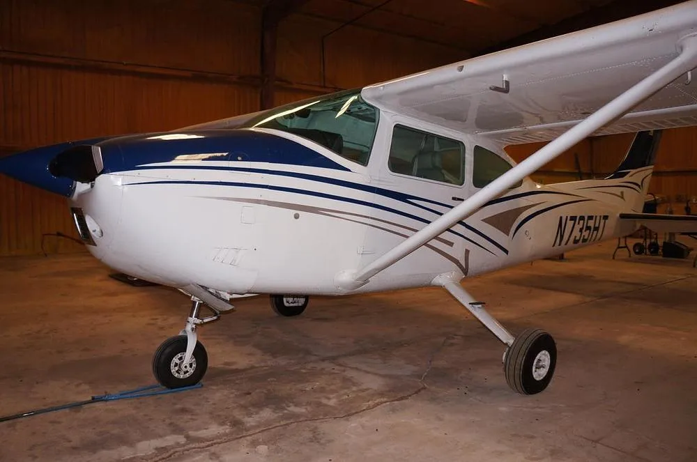A white and blue Cessna 182 Skylane N735HT is parked in a hangar, viewed from the front-left.