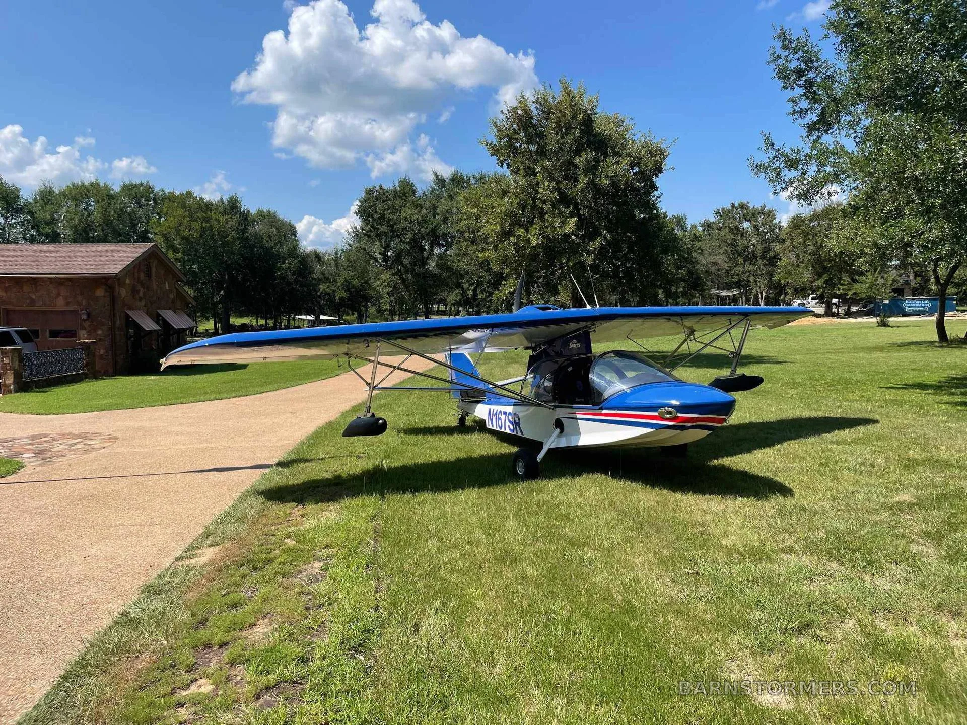 A blue and white Searey amphibian aircraft with registration N167SR parked on grass next to a gravel path and a building.