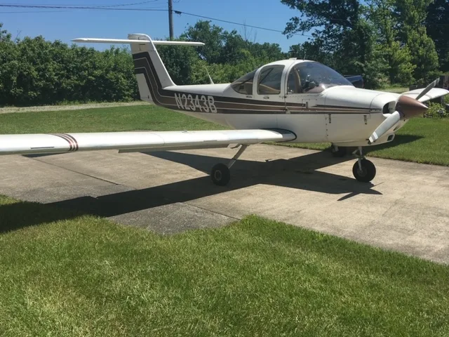 A Mooney M20C aircraft with a white and brown paint scheme is parked on a concrete surface.