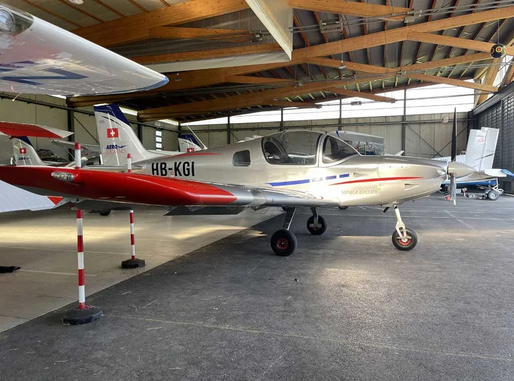 A silver Sonaca 200 aircraft with red wingtips and blue fuselage stripe, registration HB-KGI, parked inside a hangar.