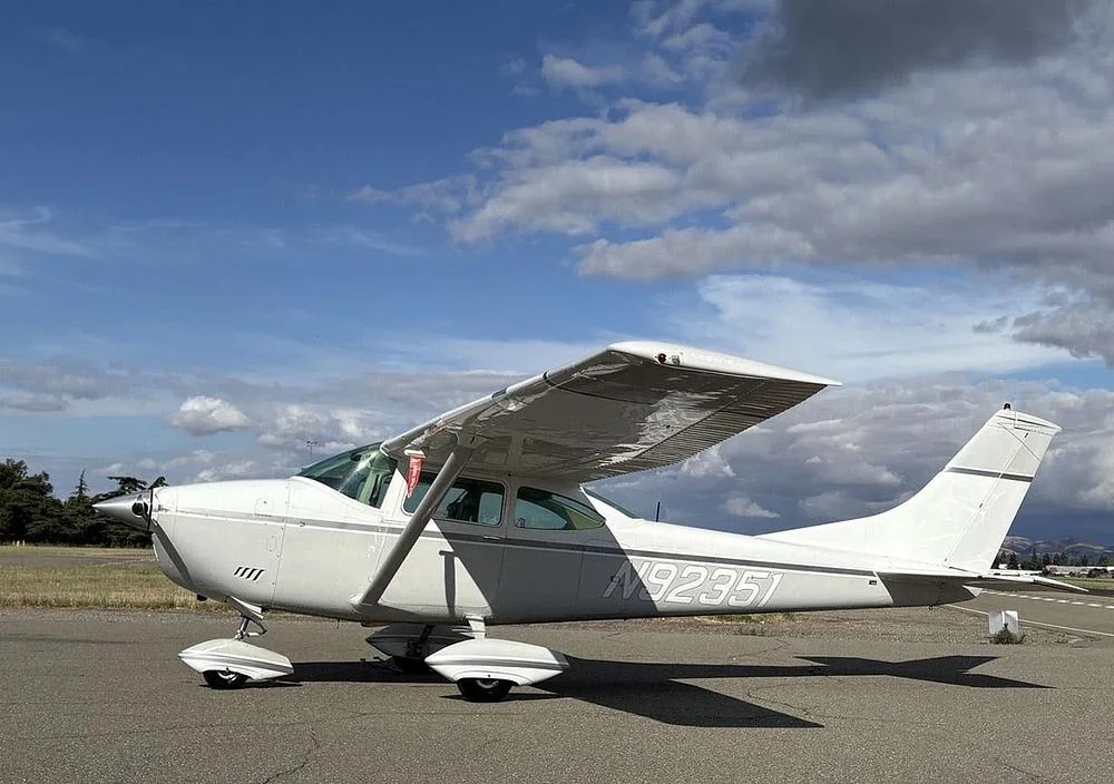 A white Cessna 172 with gray stripes, registration N92351, parked on an airfield with a cloudy sky.