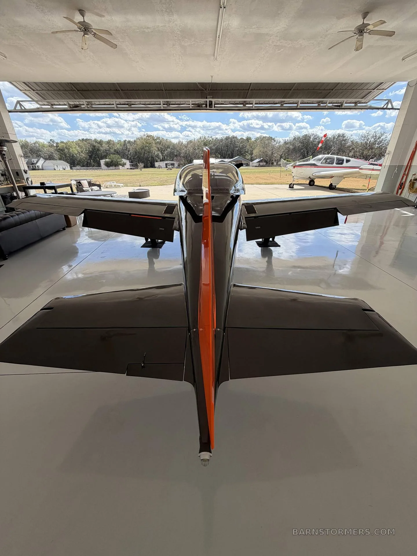 A rear view of the black and orange single-engine aircraft with a prominent orange vertical stabilizer, looking out of a hangar.