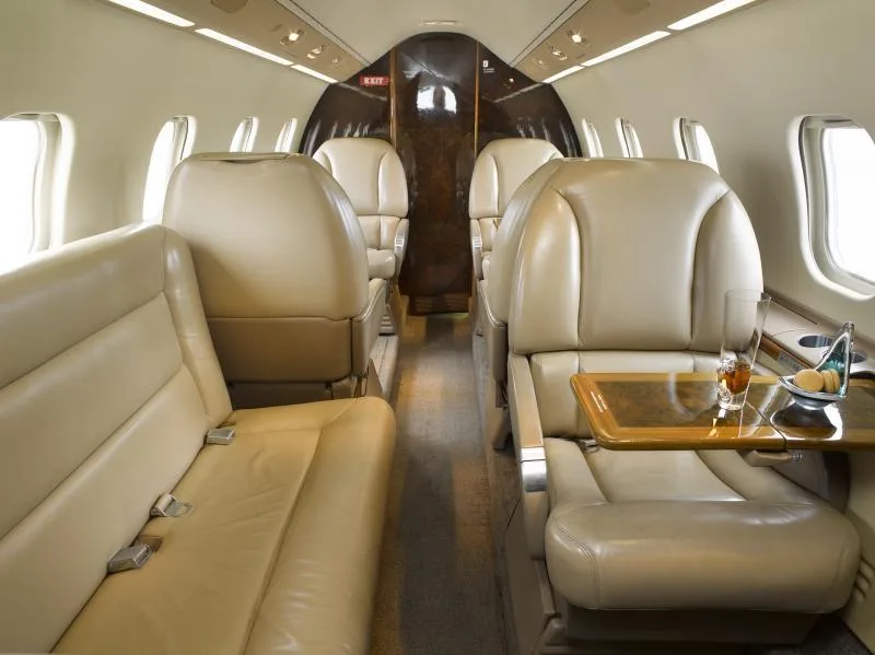 View of the aircraft cabin interior with tan leather seats, a side couch, and a polished wood table with a drink and snacks.