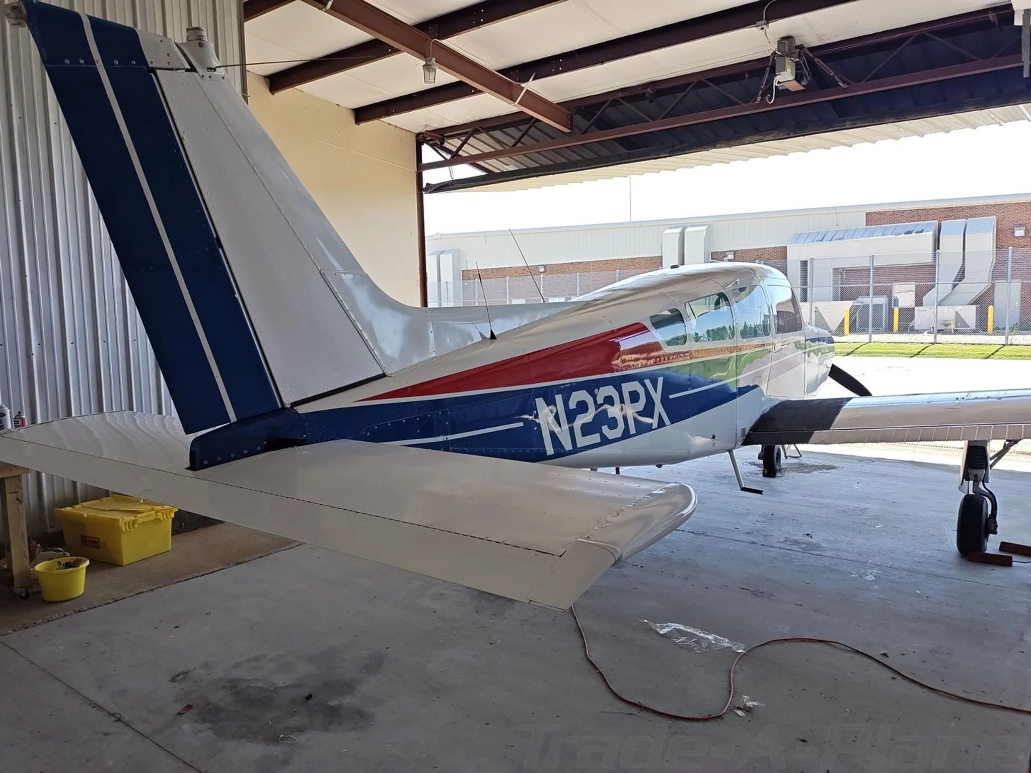 A white Beechcraft Sierra N23PX with blue and red stripes parked in a hangar, showing the tail and right side.