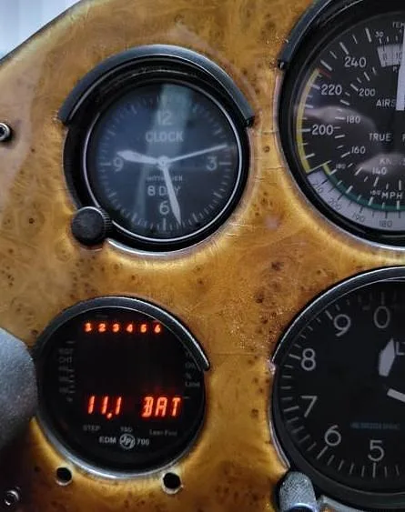 A close-up of the aircraft's wood grain instrument panel, showing a clock and a digital battery display.