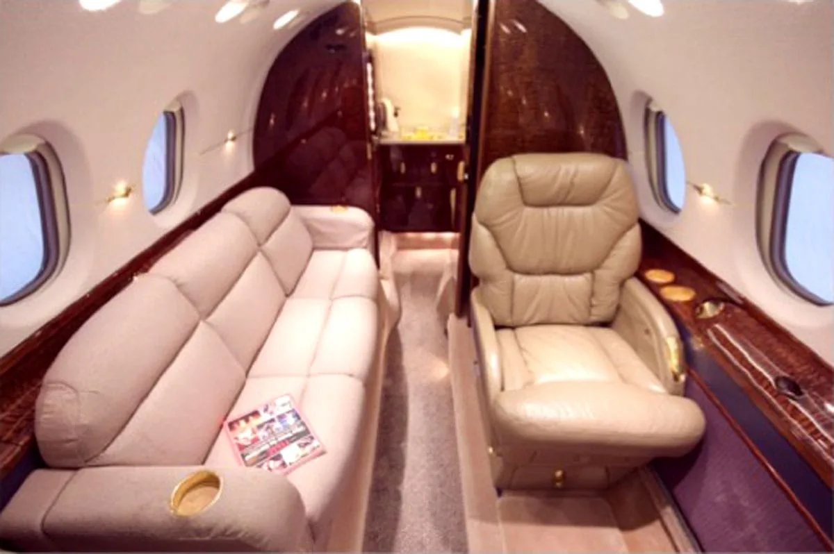 The luxurious aircraft cabin features a beige sofa on the left and a single tan leather seat on the right, with dark wood accents.