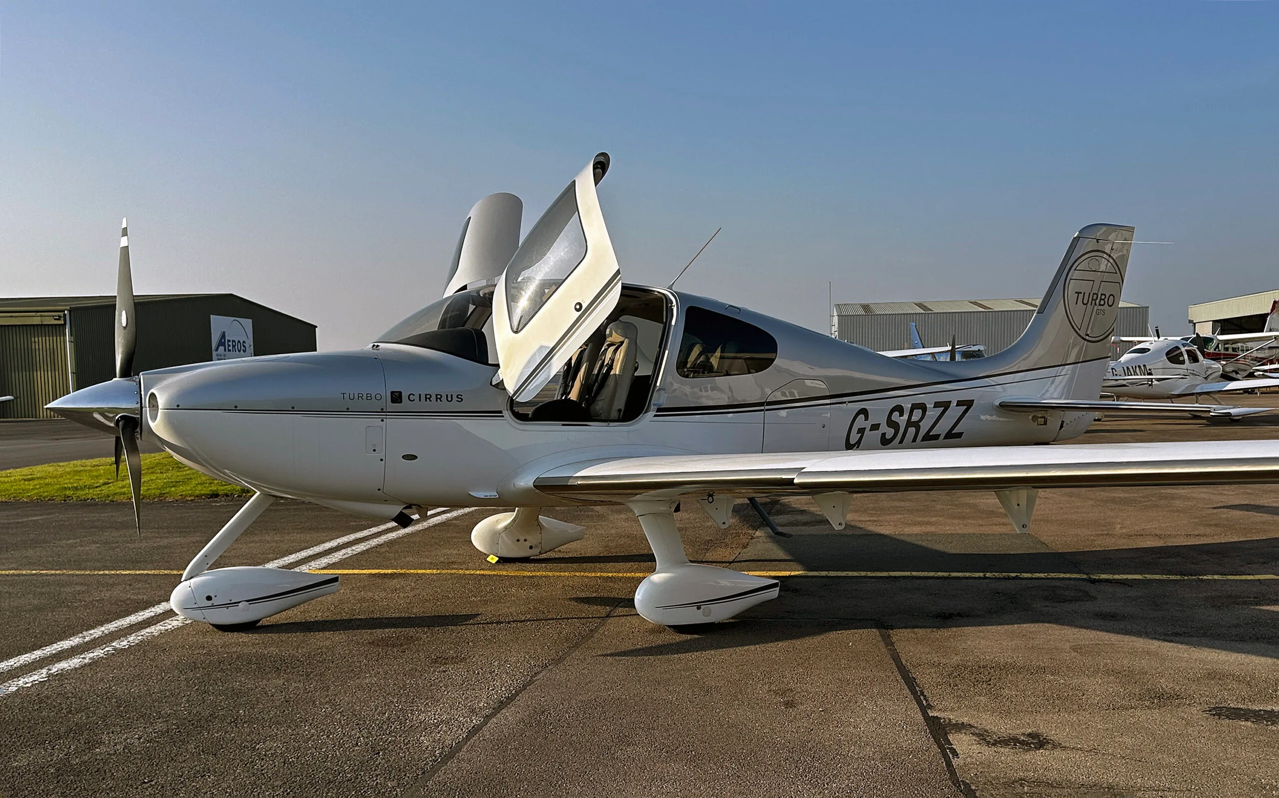 A silver and white Cirrus SR22 Turbo aircraft with registration G-SRZZ and open doors, parked on an airfield.
