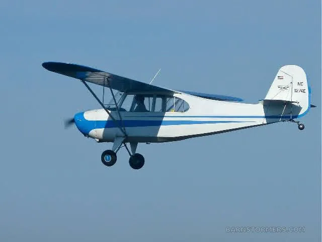A white and blue single-engine high-wing aircraft, registration NC 1274E, flying in a clear blue sky.