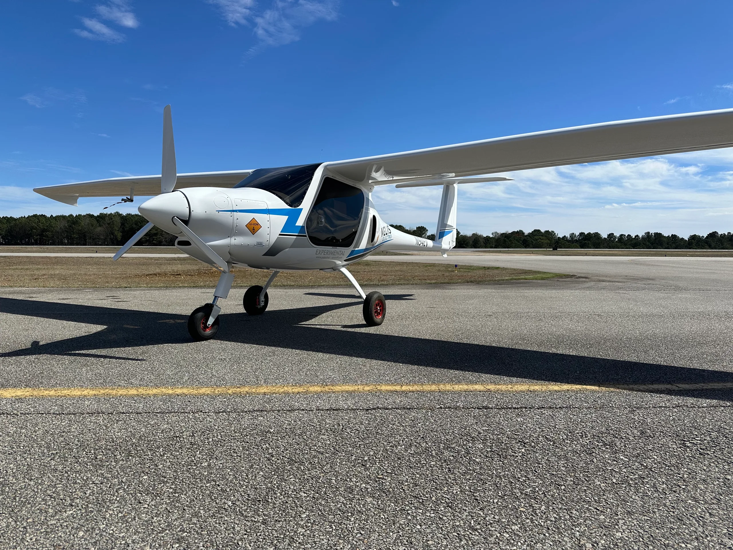 A white Pipistrel Velis Electro aircraft with blue and grey accents is parked on an asphalt tarmac under a clear blue sky.