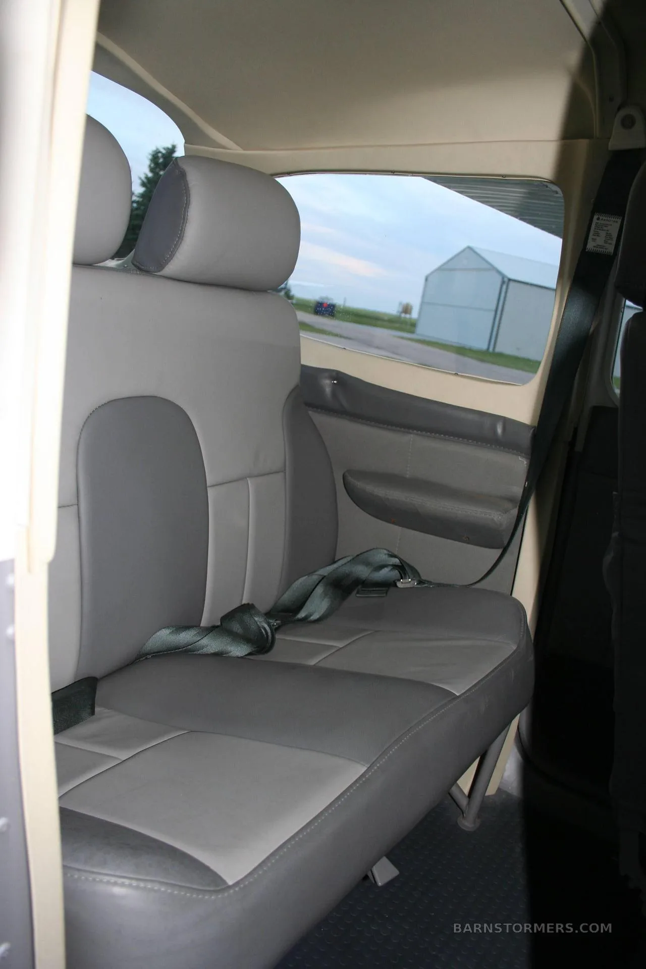 The gray and light gray rear seats of a small aircraft cabin are visible, with a view of a hangar outside.