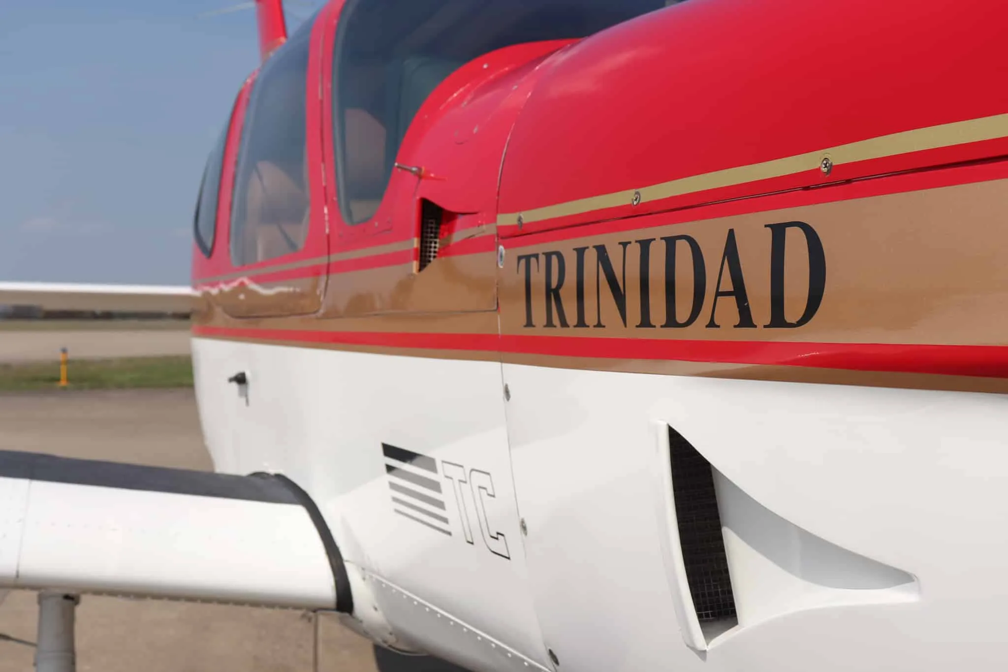 A close-up view of the fuselage of a red and white Socata Trinidad aircraft, showing the 'TRINIDAD' lettering and gold stripe.