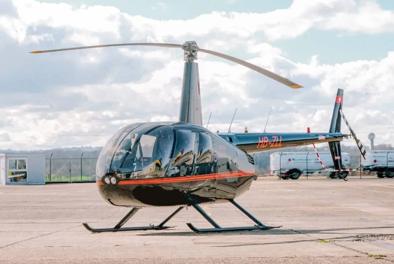 A black Robinson R44 Raven II helicopter with orange stripes, registration HB-ZLL, parked on an airfield under a cloudy sky.