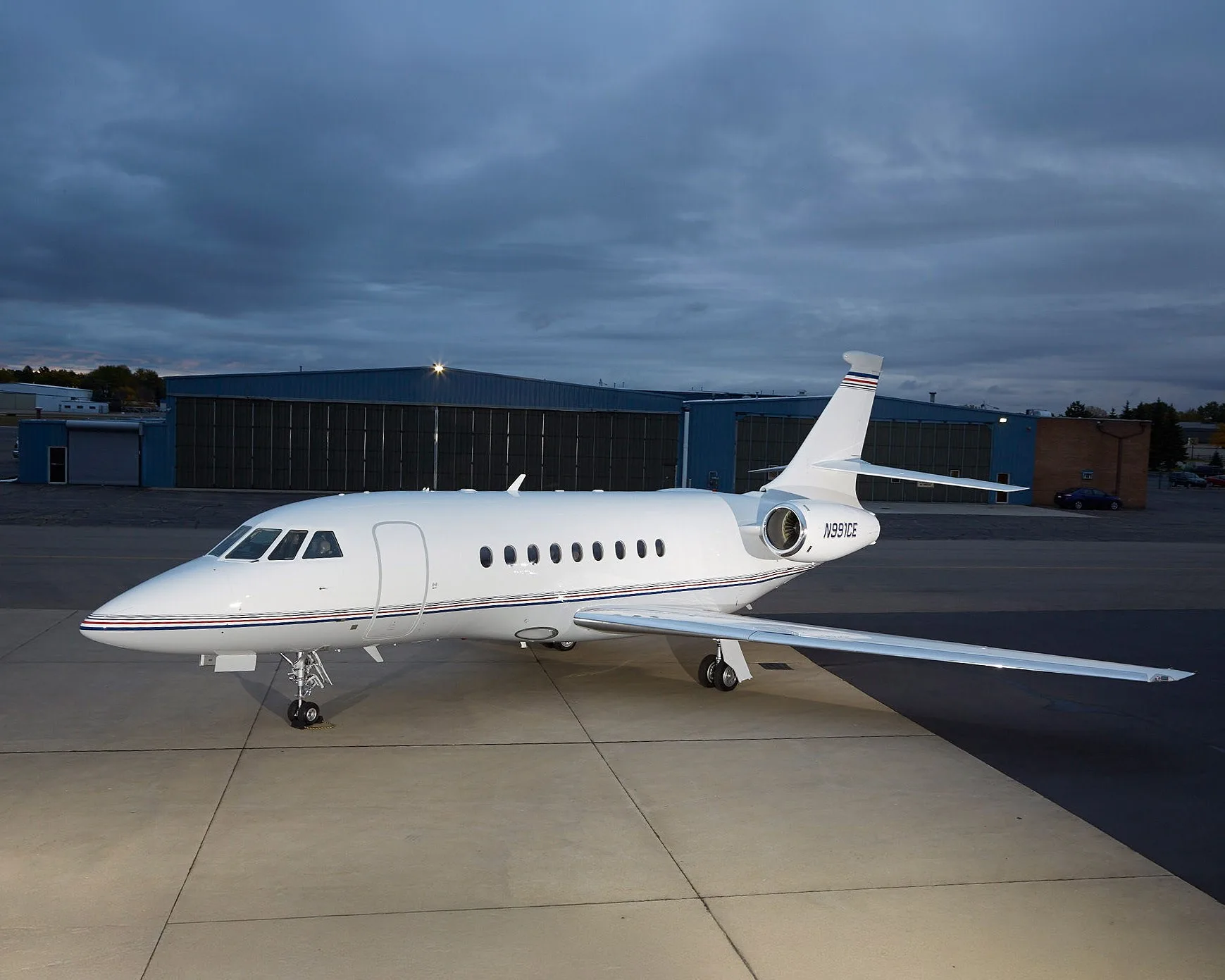 A white Dassault Falcon 900EX aircraft with blue and red stripes on the tail and fuselage, parked on an tarmac at dusk with hangars in the background.