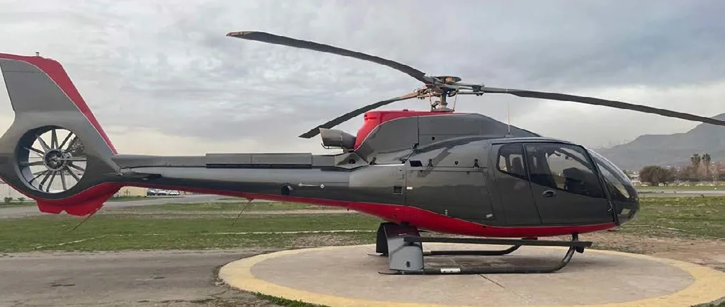 A dark grey and red helicopter is parked on a circular helipad with a grassy field and mountains in the background.