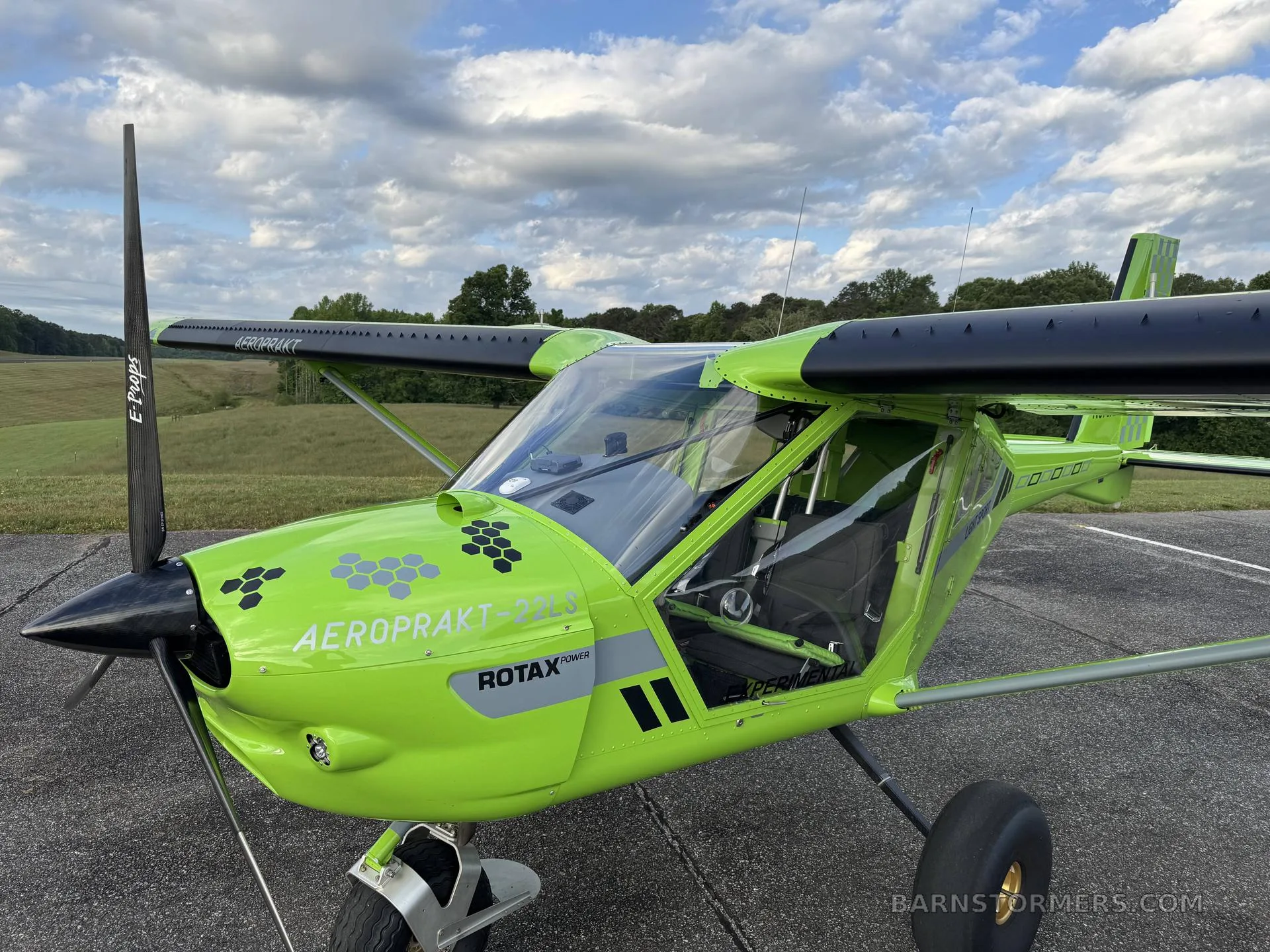 A three-quarter front view of a bright green Aeroprakt-22LS light sport aircraft with black wings and a clear canopy, parked on an asphalt runway.