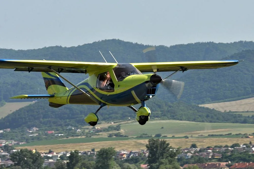 A yellow and blue Skyper GT9 aircraft flying low over a town with green hills in the background.