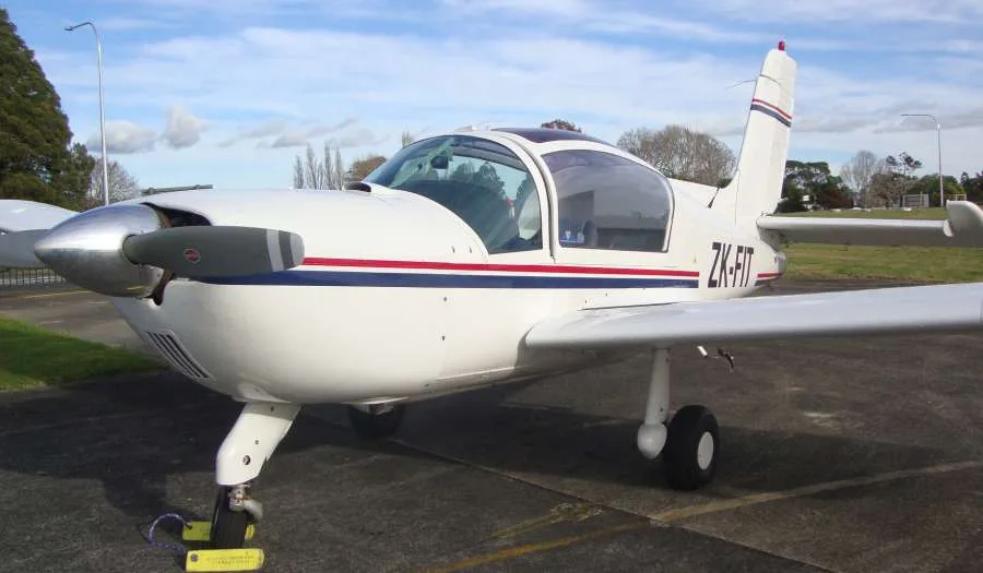 A white single-engine propeller aircraft with blue and red stripes, registration ZK-FIT, parked on an airfield tarmac.
