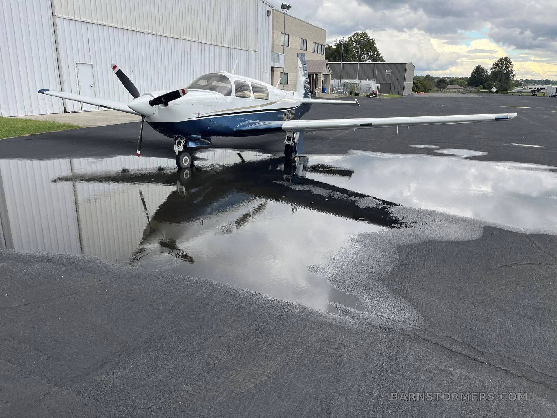 A white and blue single-engine propeller aircraft parked on an asphalt ramp next to a hangar, reflected in a large puddle under a cloudy sky.