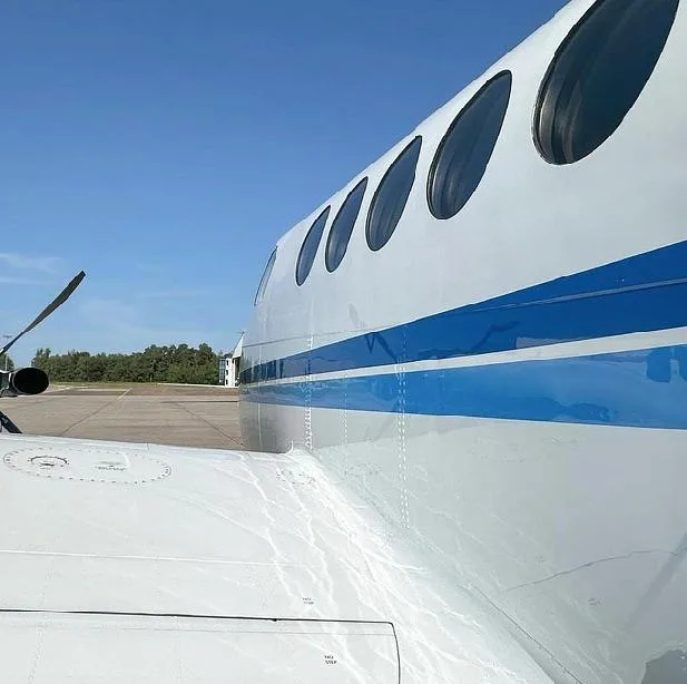 A close-up view of the white fuselage with blue stripes and windows, and the wing of a twin-turboprop aircraft.