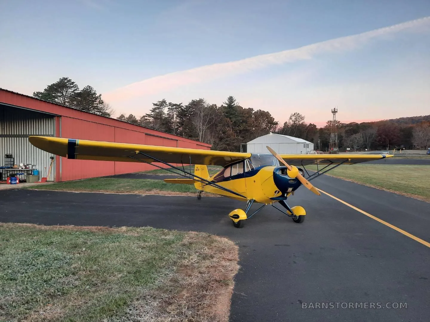 A yellow and blue Aeronca Champ aircraft is parked on an asphalt ramp next to a red hangar at sunset.