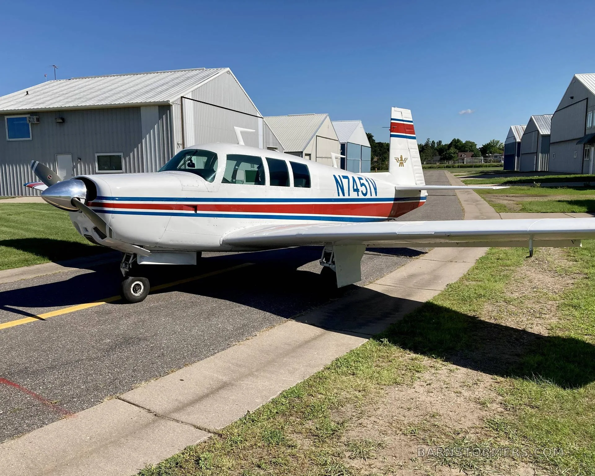 A white single-engine aircraft with red and blue stripes, registration N7451V, parked on a tarmac in front of hangars on a sunny day.