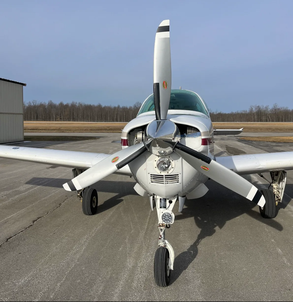 A head-on view of a white and maroon Beechcraft Bonanza aircraft with its propeller centered on a tarmac.