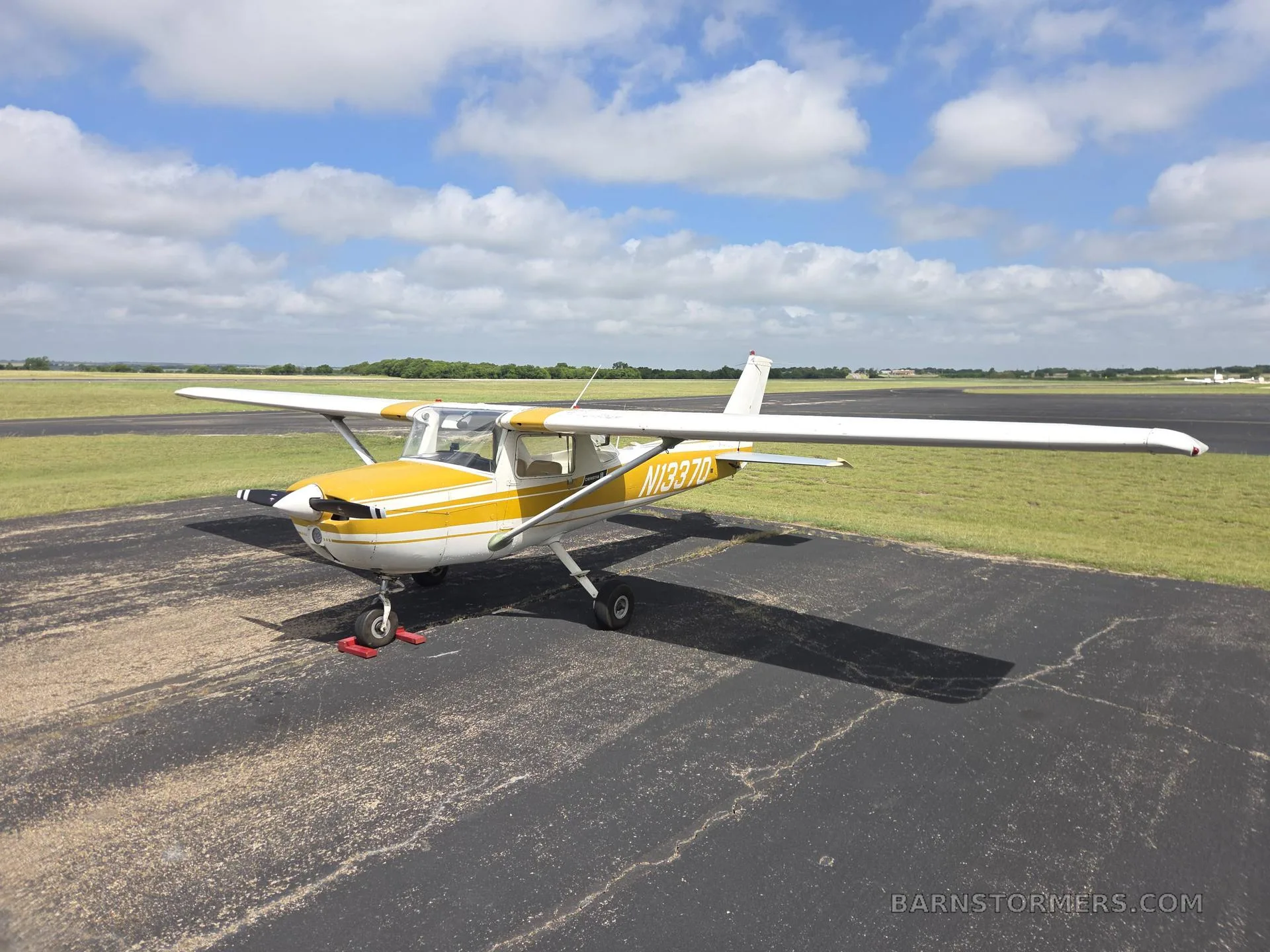 A white and yellow Cessna 150 or similar light aircraft (N13370) parked on an asphalt ramp under a partly cloudy sky, viewed from the front-left.