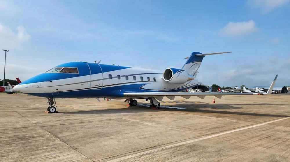 A blue and white Bombardier Challenger 604 private jet is parked on a sunny airport tarmac.