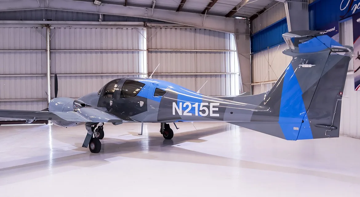 A slightly different side view of a gray and blue Diamond DA62 aircraft, registration N215E, in a hangar.