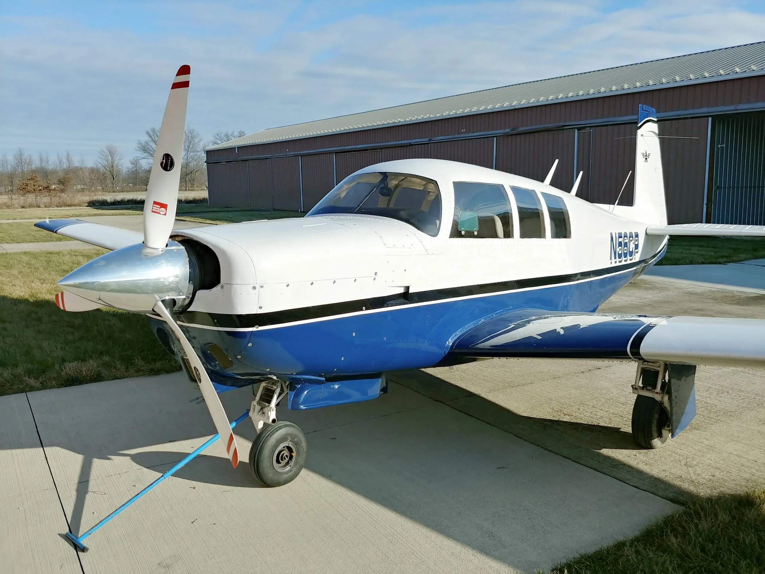 A blue and white Mooney aircraft, registration N56CP, is parked on the tarmac in front of a hangar.