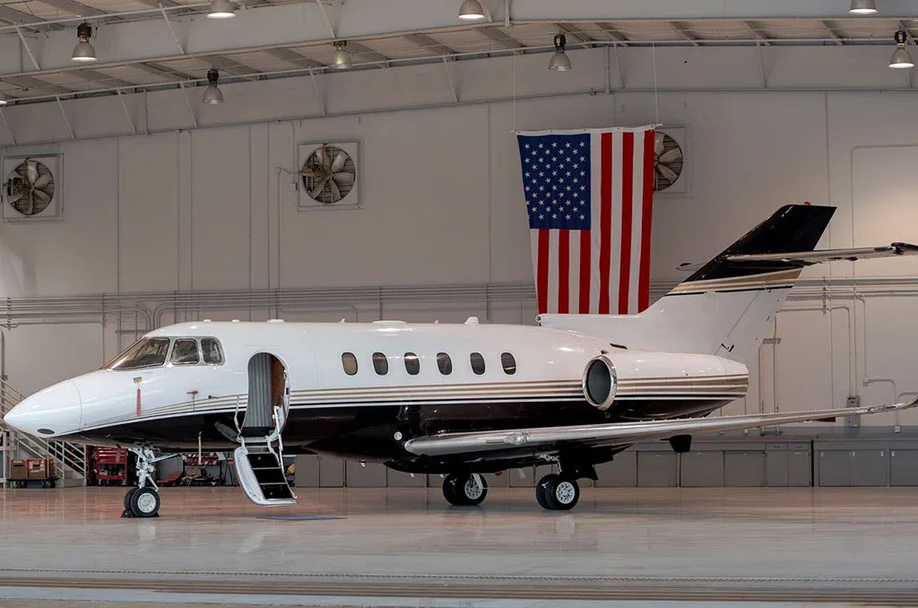 A white and black private jet with its main cabin door open and stairs extended, parked in a brightly lit hangar with an American flag.