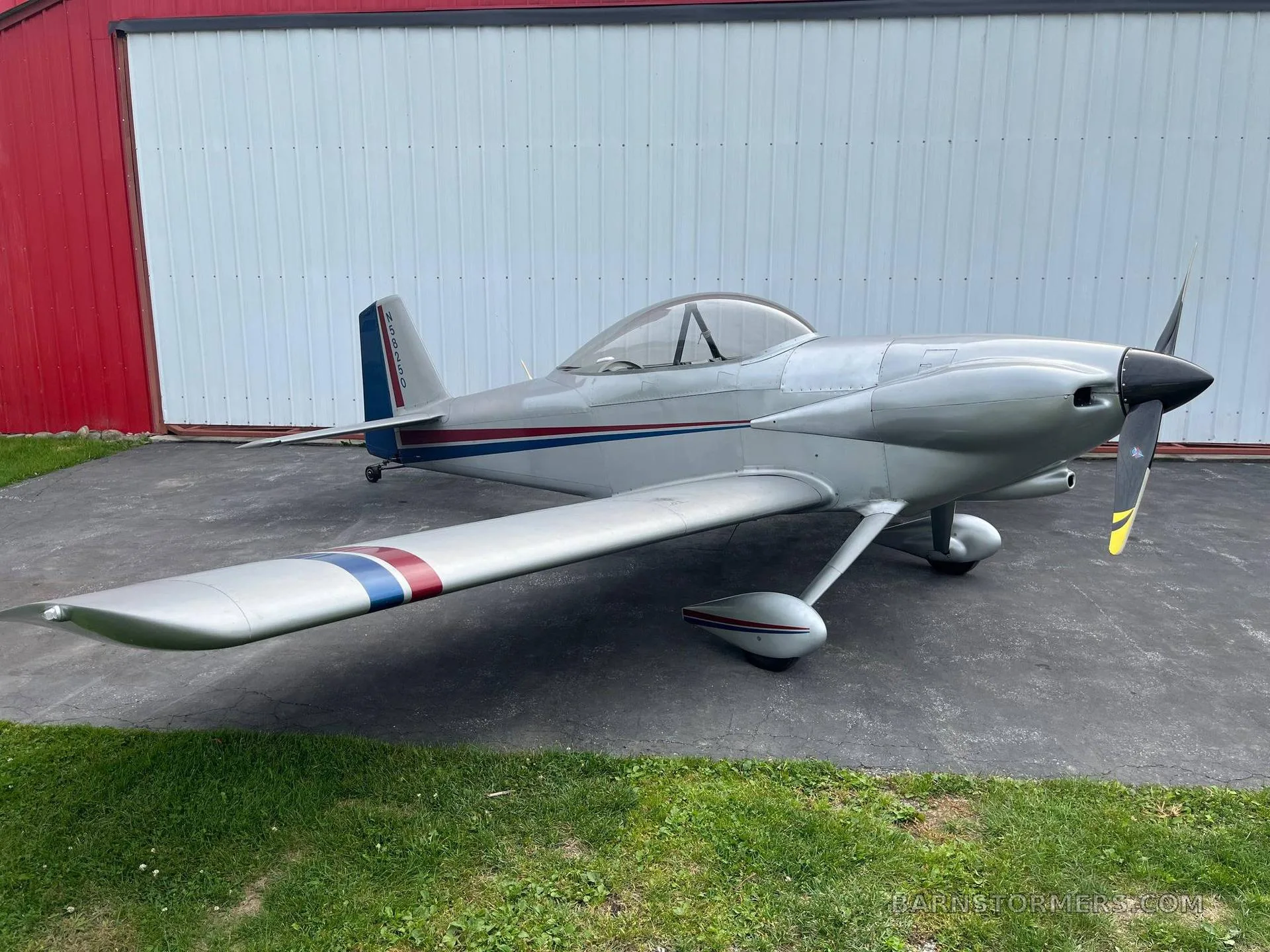 A silver single-engine aircraft with red and blue stripes on the tail and wings, parked on asphalt next to a white building.